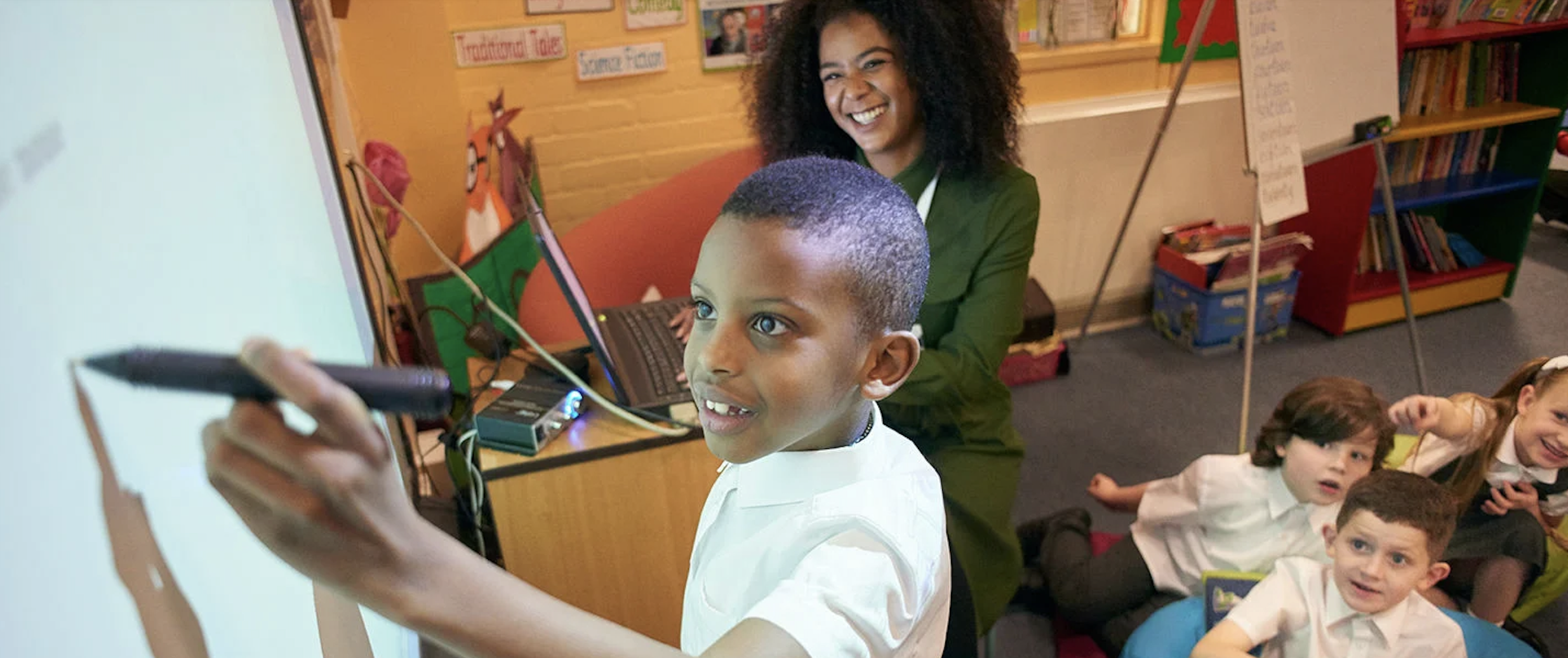 Student writing on a smart board in front of the classroom while his teacher and classmates smile and watch.