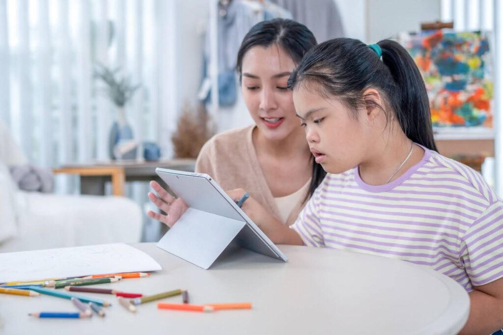 Disabled student sitting at a table is assisted while using a tablet.